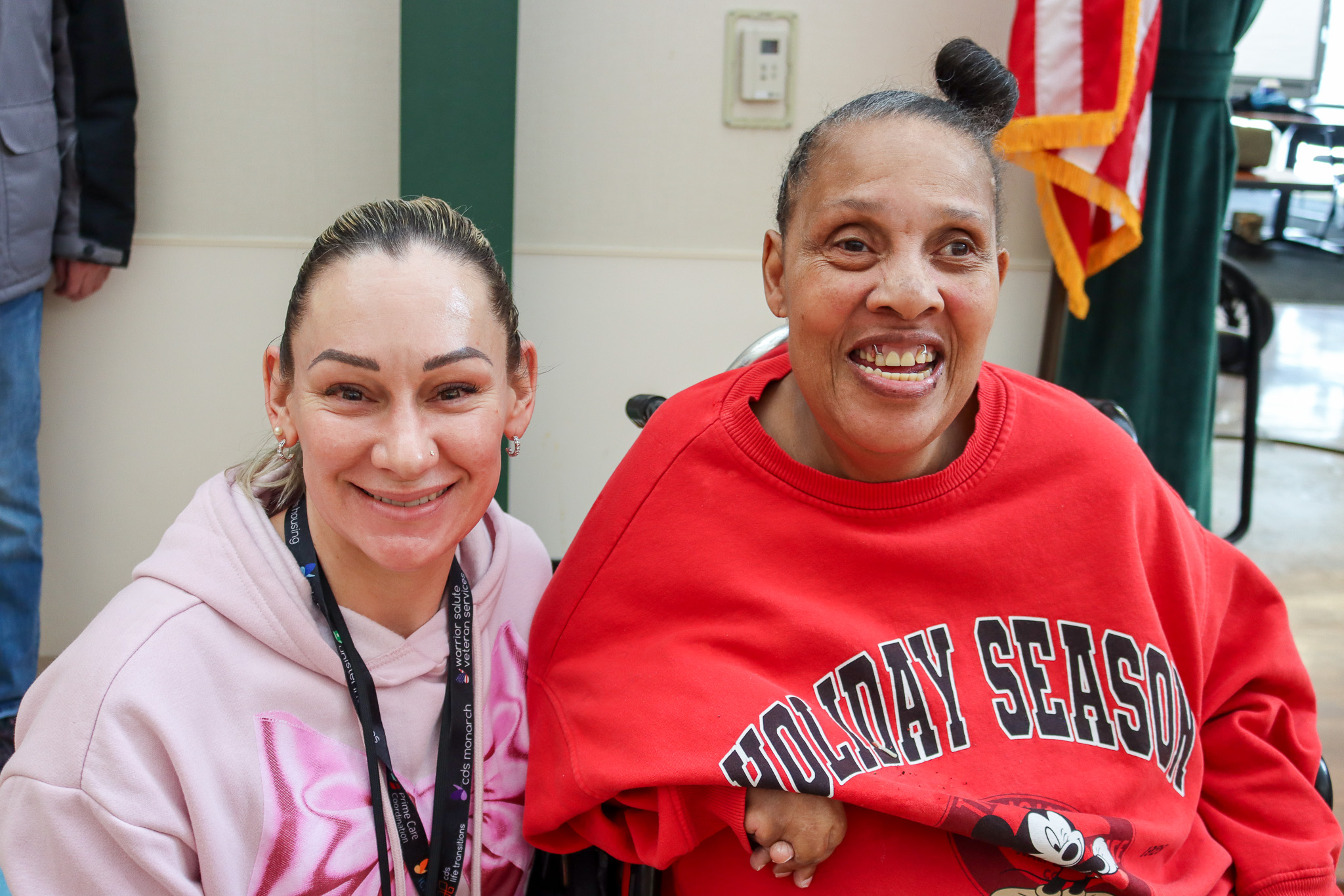 Two smiling women indoors. One is wearing a red “Holiday Season” Mickey Mouse sweatshirt and sitting in a wheelchair, while the other wears a pink hoodie and sits beside her.