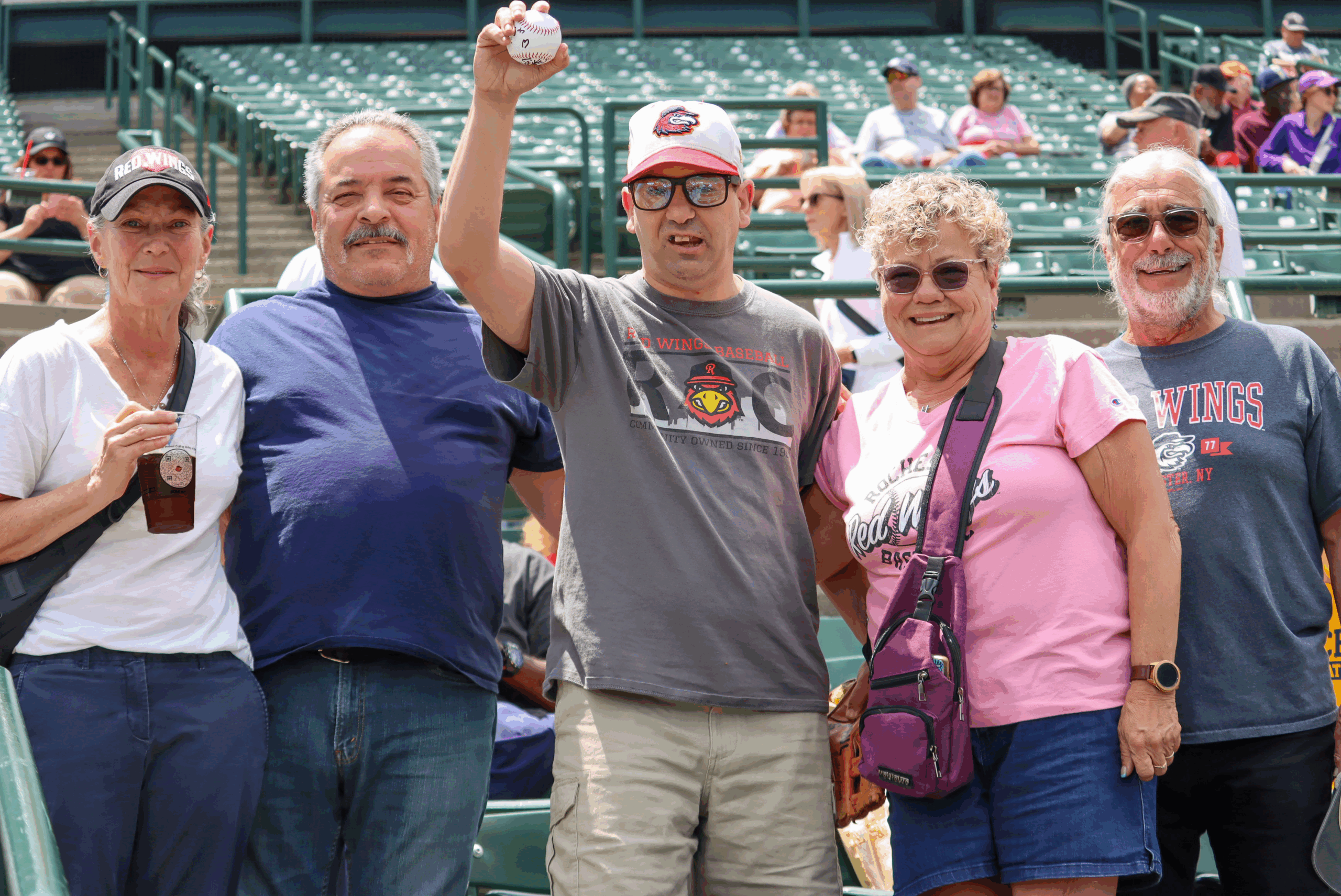 A group of five people smiling at a baseball stadium. One man holds up a baseball in the center. The stadium seating is visible in the background.