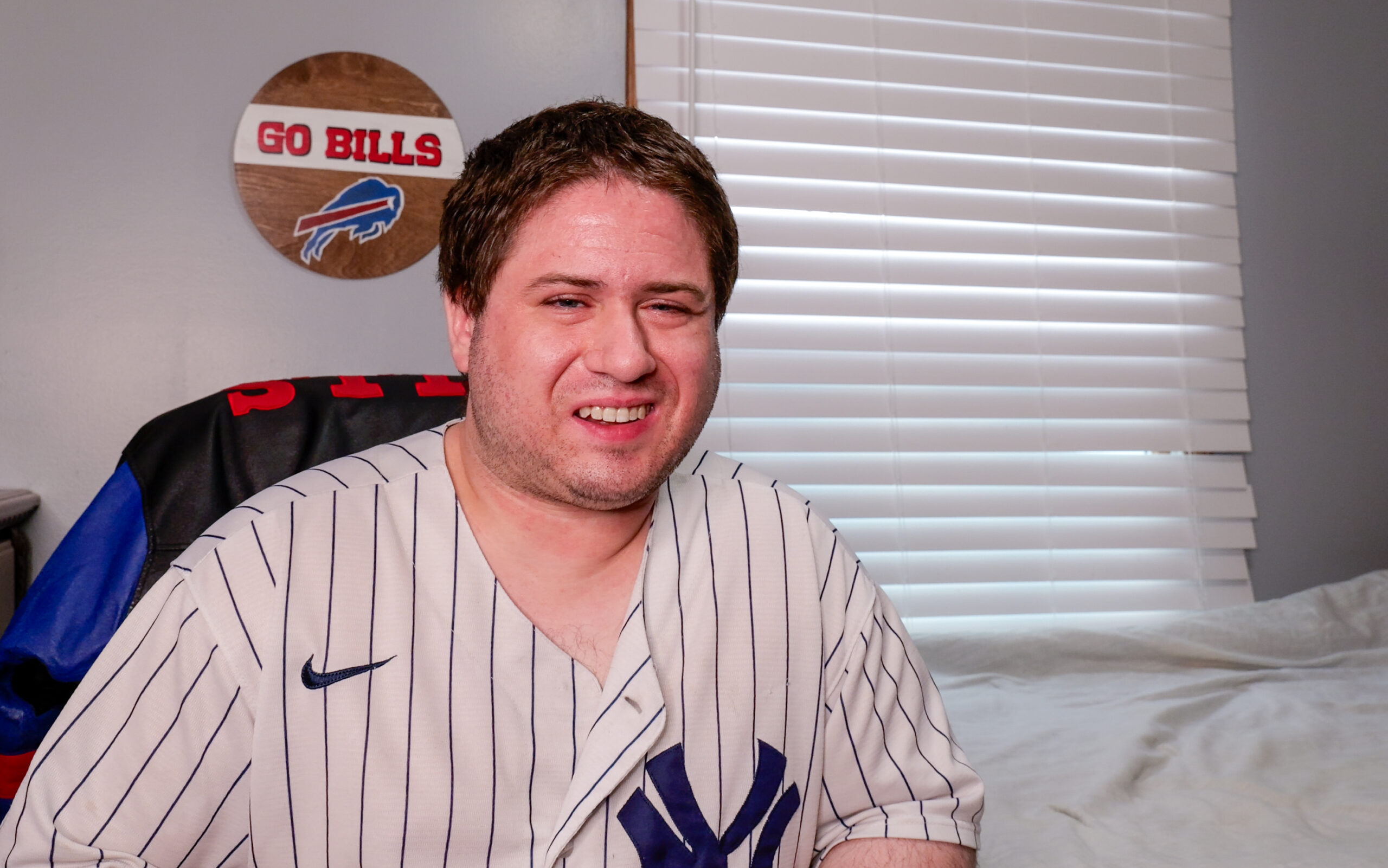 A man wearing a New York Yankees jersey sits in front of a wall with a “Go Bills” sign. He is smiling and seated in a comfortable chair with blinds behind him.
