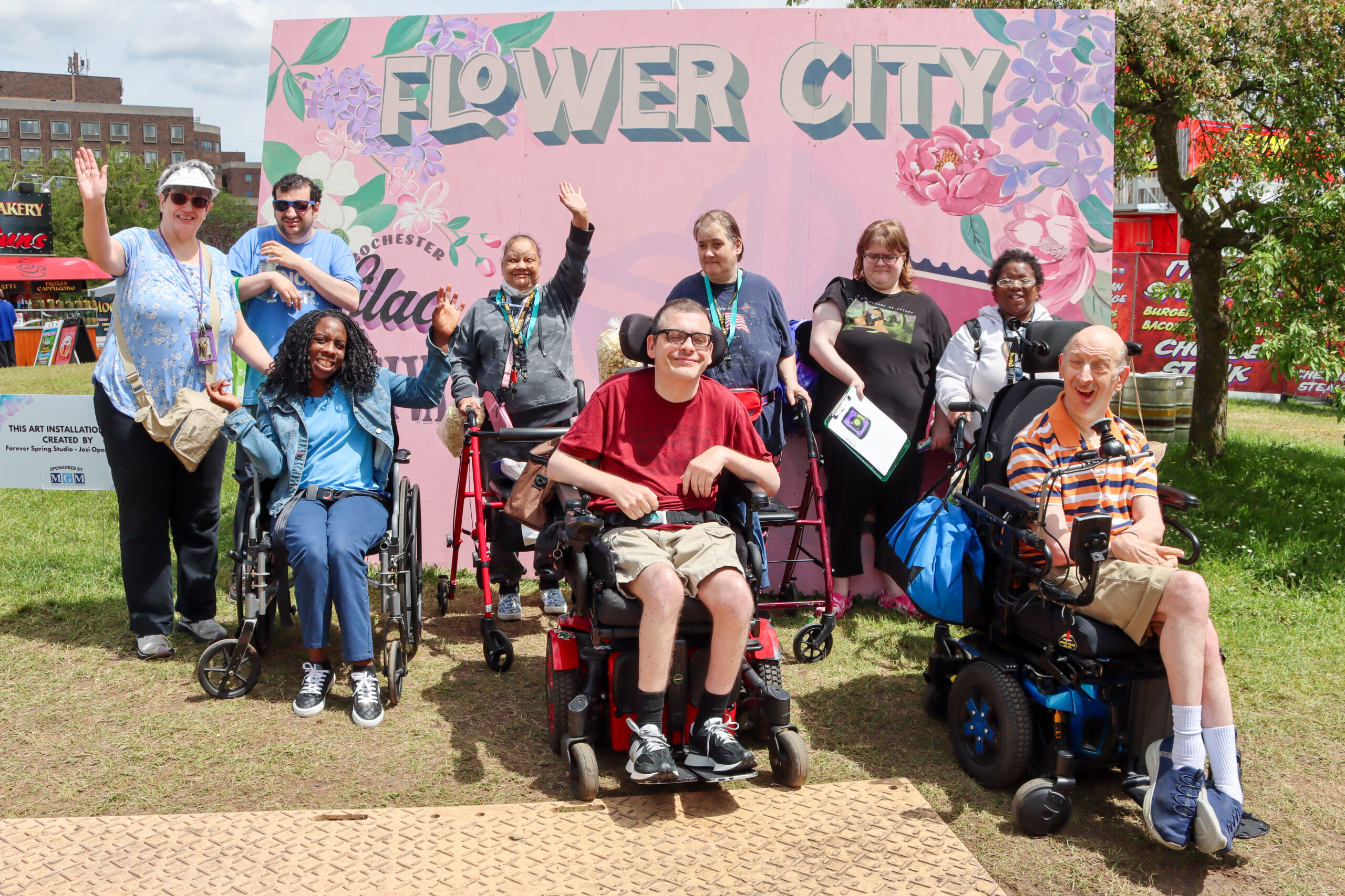 Group of individuals posing and smiling at an outdoor event in front of a “Flower City” mural. Several people are using wheelchairs, and others stand around them.